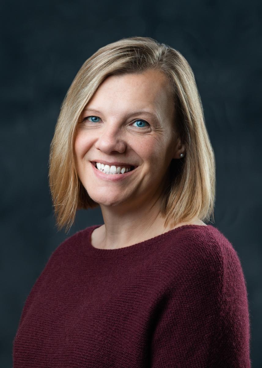 Headshot of a middle-aged, blond-haired, fair-skinned female wearing a maroon sweater an smiling, but only slightly
